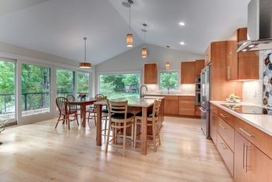 Open kitchen and dining area with maple cabinets, a large island, vaulted ceiling and a wall of glass facing the trees in Lexington, MA
