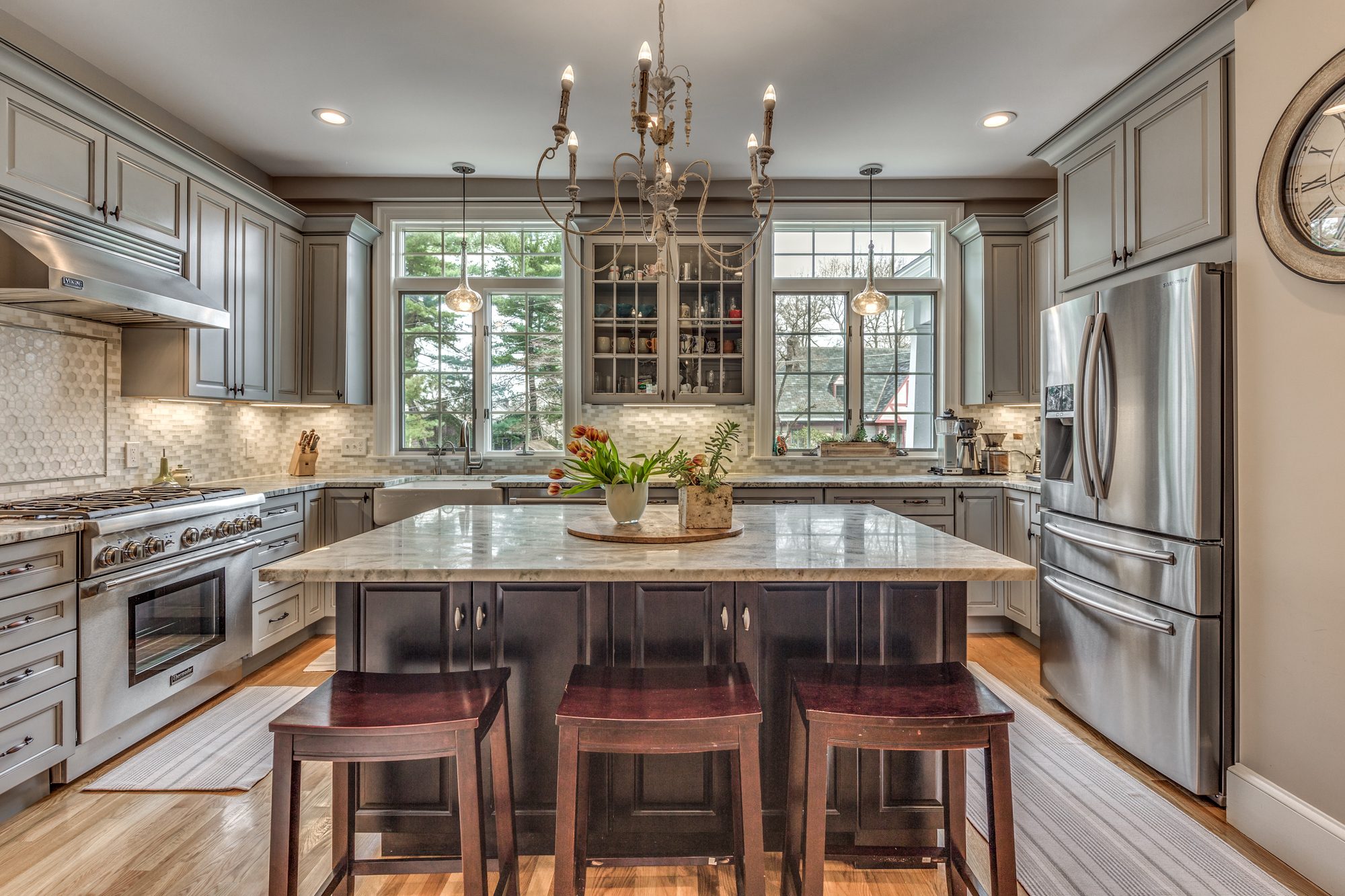 Kitchen with gray painted cabinets, a dark cherry island with stools, a stone top and a pro-style range under a chandelier, in Winchester, MA