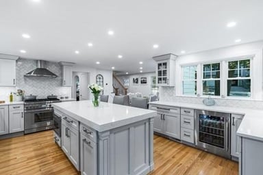 Open kitchen in Winchester, MA with gray shaker cabinets, a large white quartz island, herringbone backsplash and oak floors, with the stair beyond