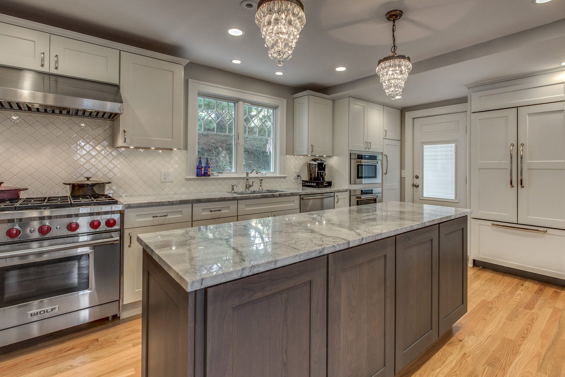 After: Sink wall: dark cabinets and black counters replaced by white Shaker cabinets, quartz counters, a pro range and hood, tile backsplash and a new leaded-glass window.