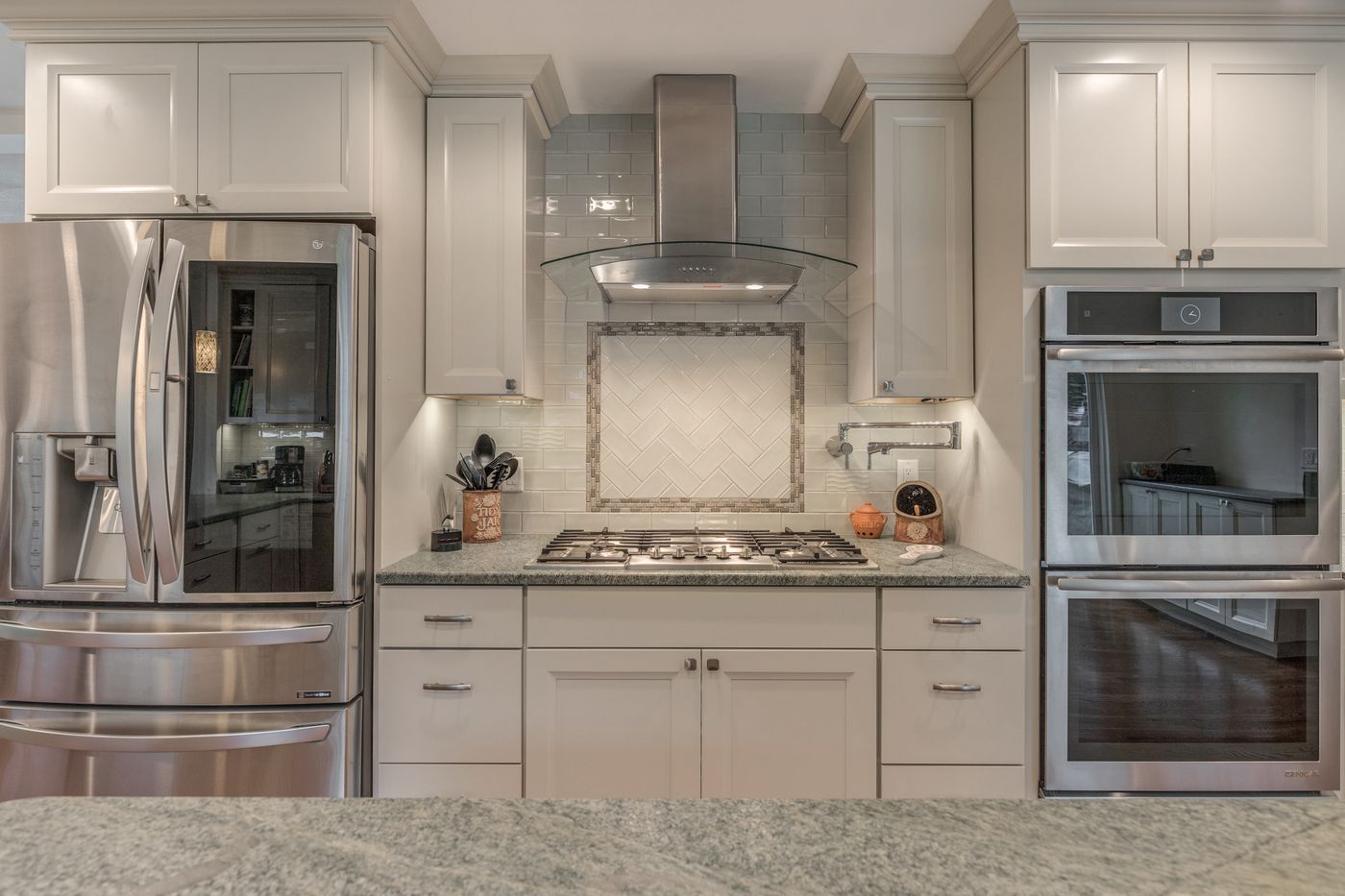 After: Cooking wall: a painted brick wall with an arched cooktop alcove and black built-in ovens became a cooktop under a glass chimney hood, a herringbone tile panel and stainless double wall ovens.