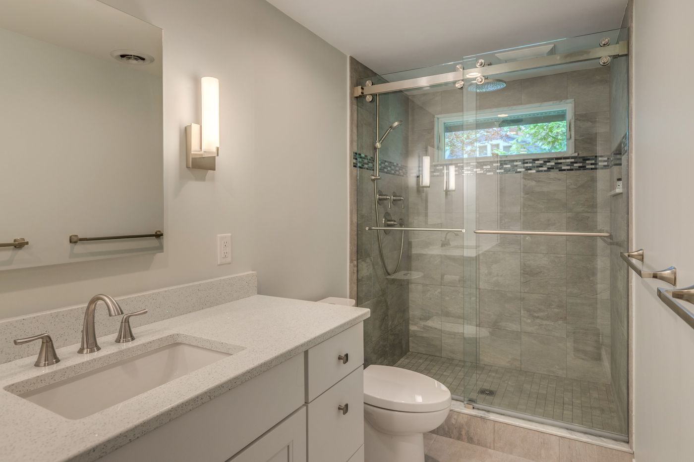 After: Primary bath: the tub and shower curtain were replaced by a walk-in shower with gray tile, a glass accent band and a sliding glass door.