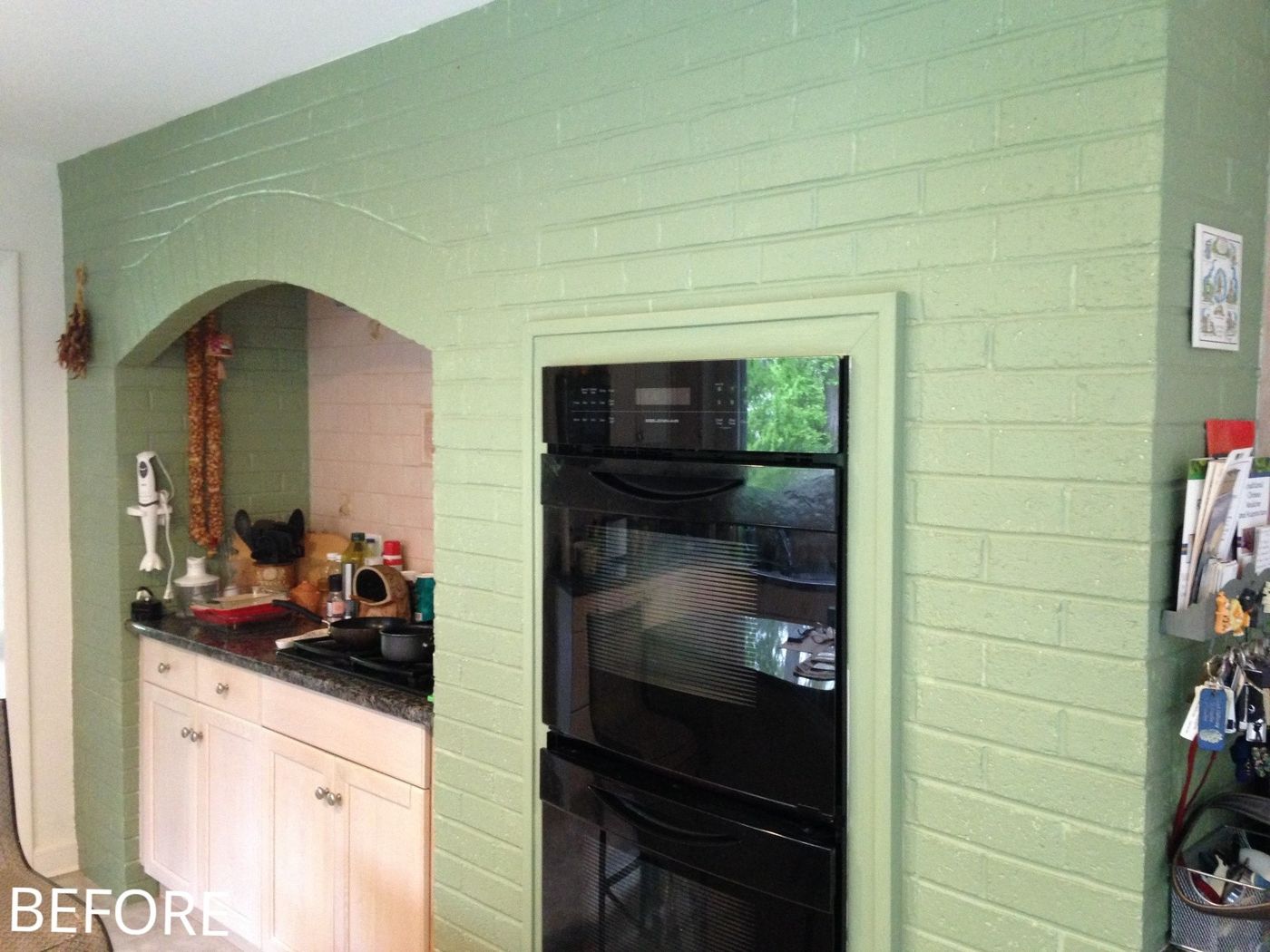 Before: Cooking wall: a painted brick wall with an arched cooktop alcove and black built-in ovens became a cooktop under a glass chimney hood, a herringbone tile panel and stainless double wall ovens.