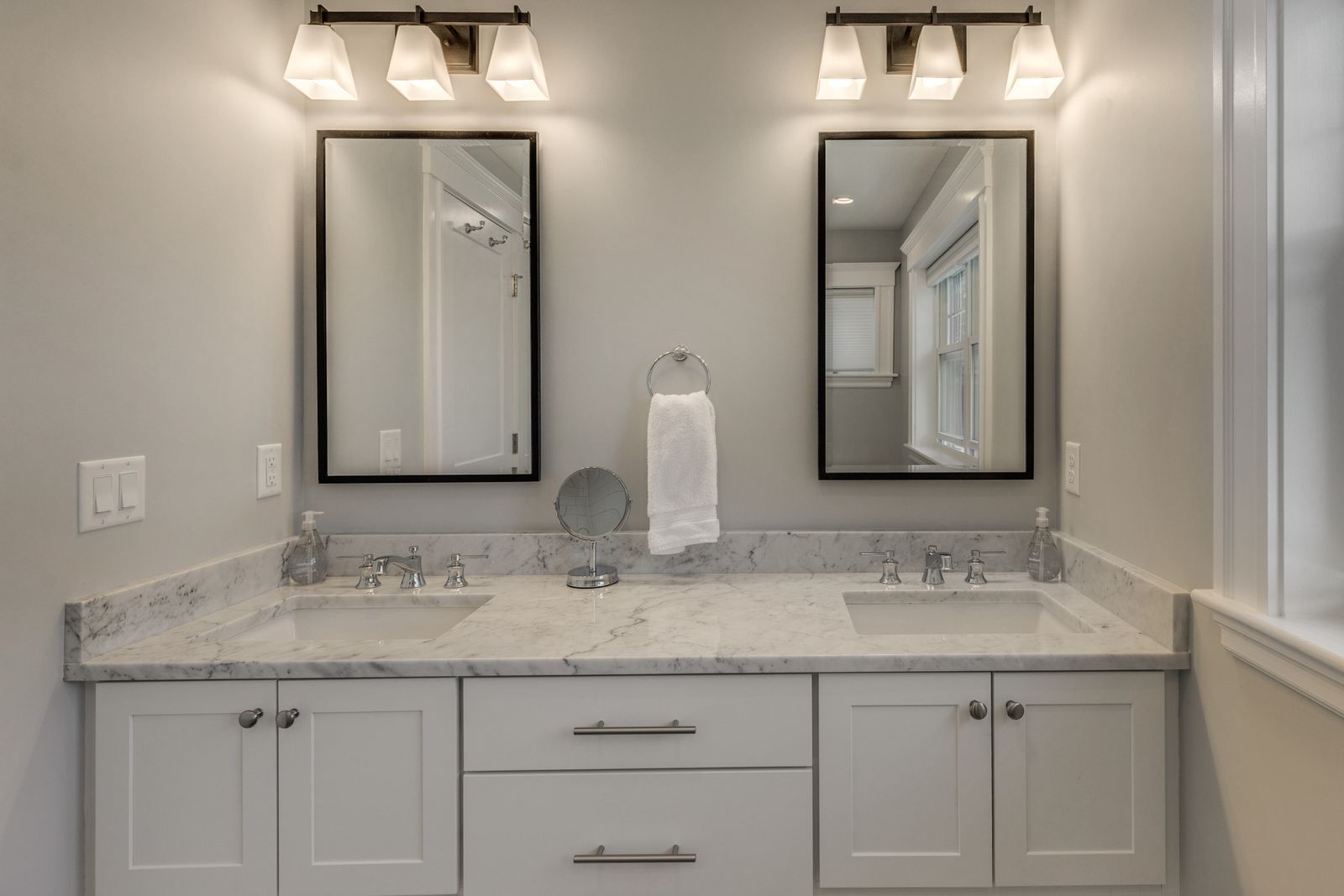White double vanity with a marble top, two black-framed mirrors and bronze light fixtures
