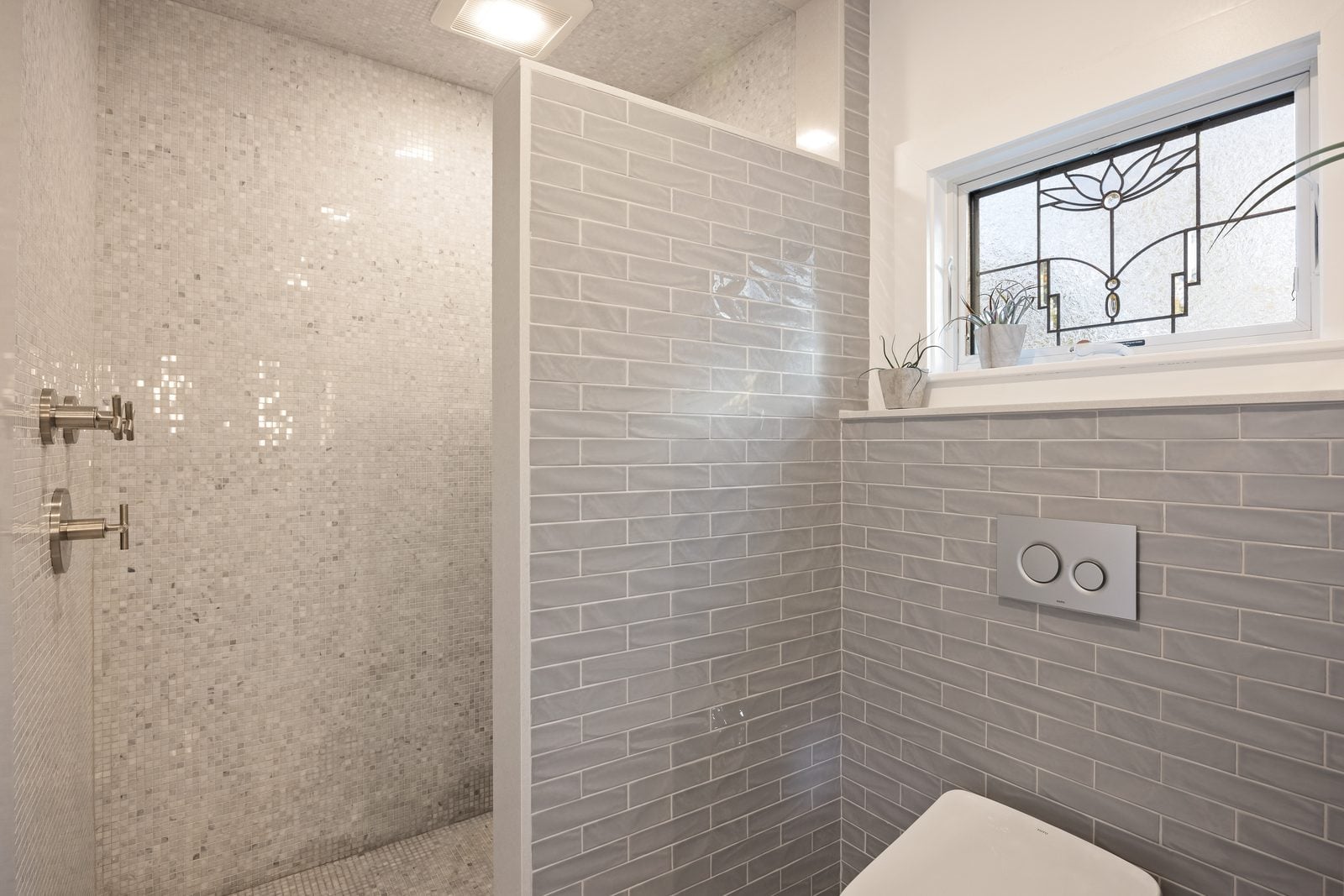 Walk-in shower in white mosaic tile beside a wall-hung toilet and leaded-glass window