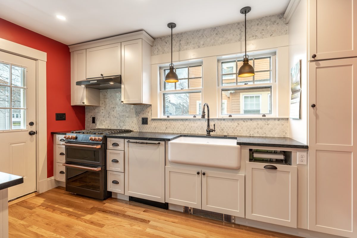 After: Kitchen sink wall: the old drainboard sink and painted cabinets became a fireclay farmhouse sink, shaker cabinets and soapstone counters under the same twin windows.