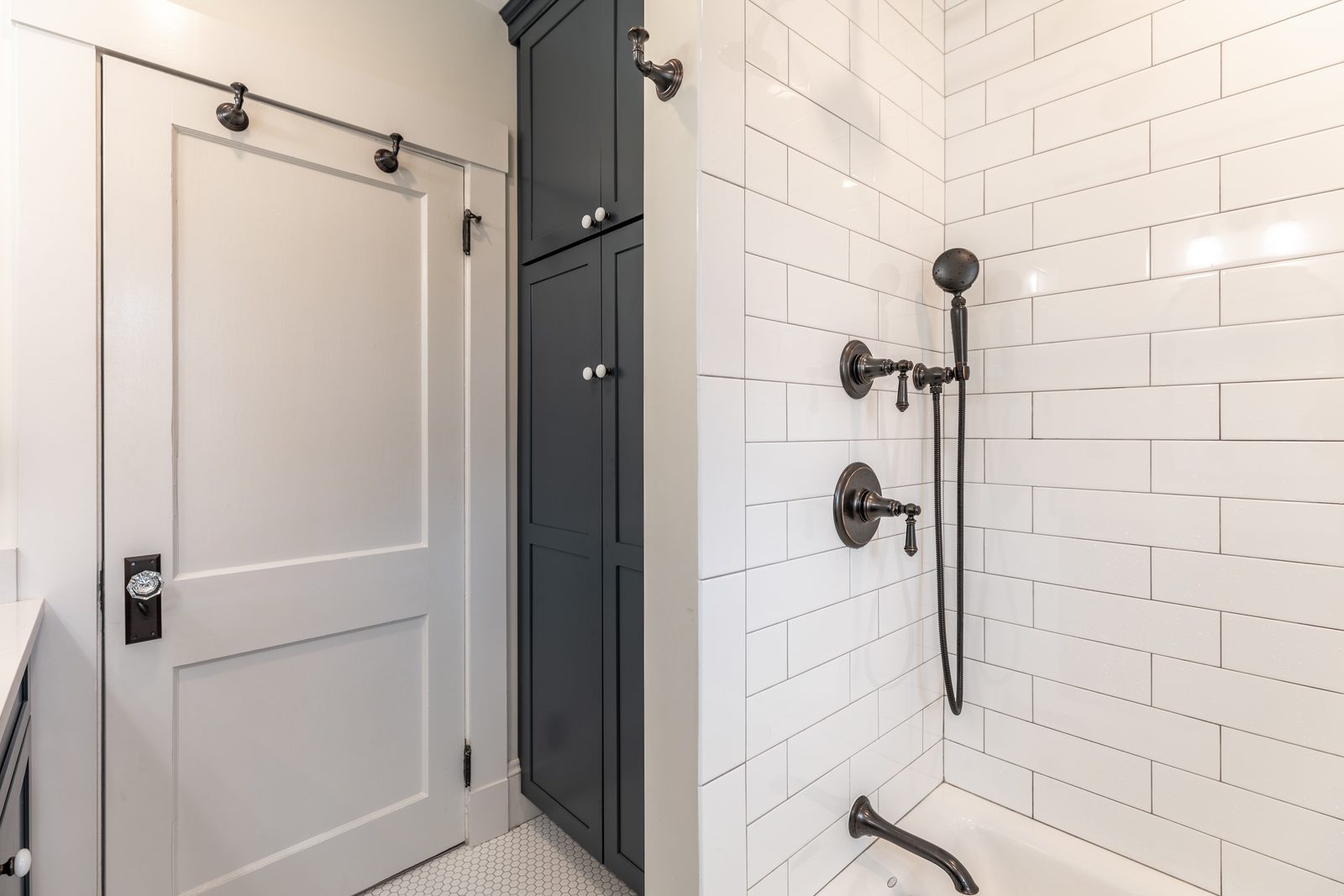 Remodeled bathroom in Melrose, MA with white subway tile tub surround, bronze shower fixtures and a tall navy linen cabinet
