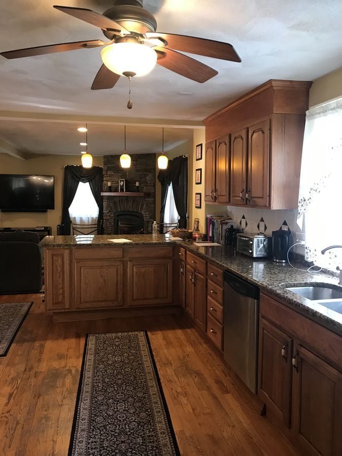 Before: Kitchen toward the family room: the oak peninsula and ceiling fan gave way to a large painted island under glass pendants, with the stone fireplace still in view.