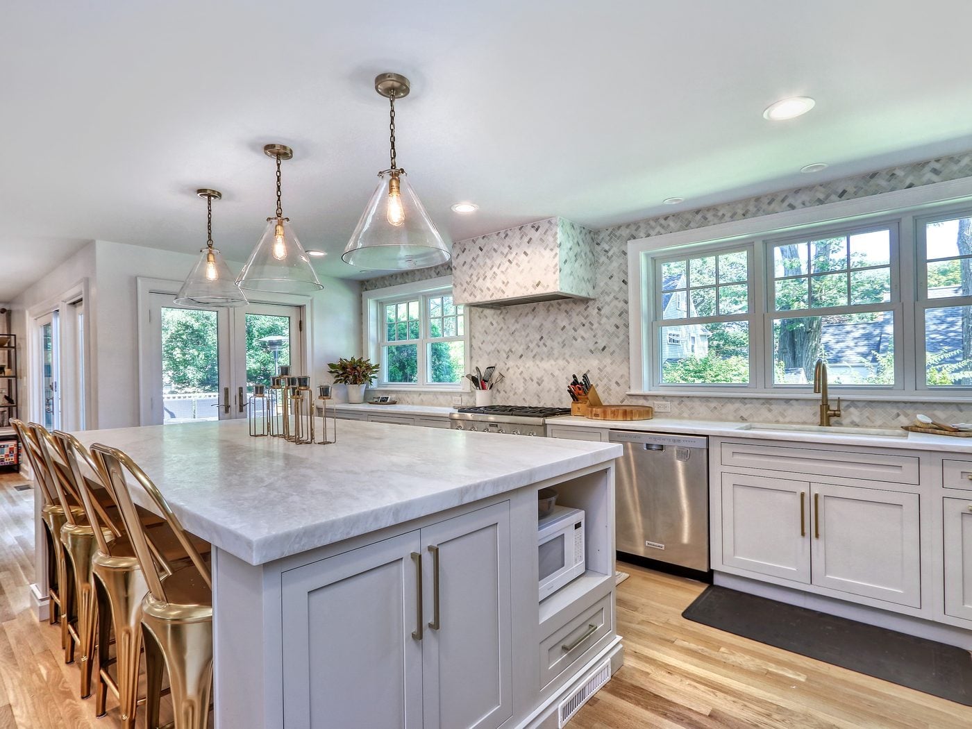 After: Kitchen: dark wood cabinets, black granite and a peninsula gave way to painted inset cabinets, a big stone-topped island and a herringbone marble wall around the same window-lit sink wall.