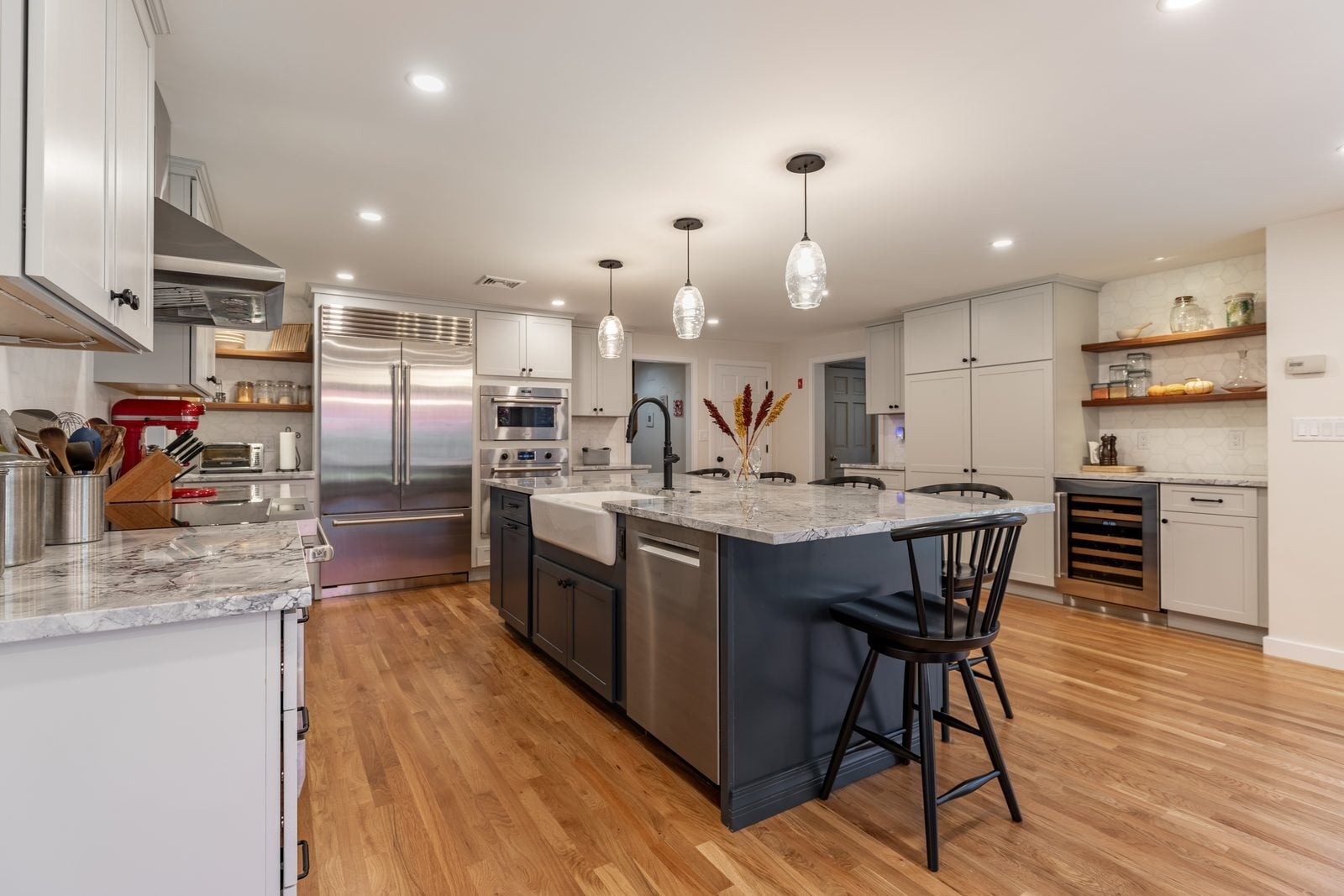 Kitchen with a built-in refrigerator, two wall ovens, floating wood shelves and a charcoal island with a farmhouse sink in Winchester, MA