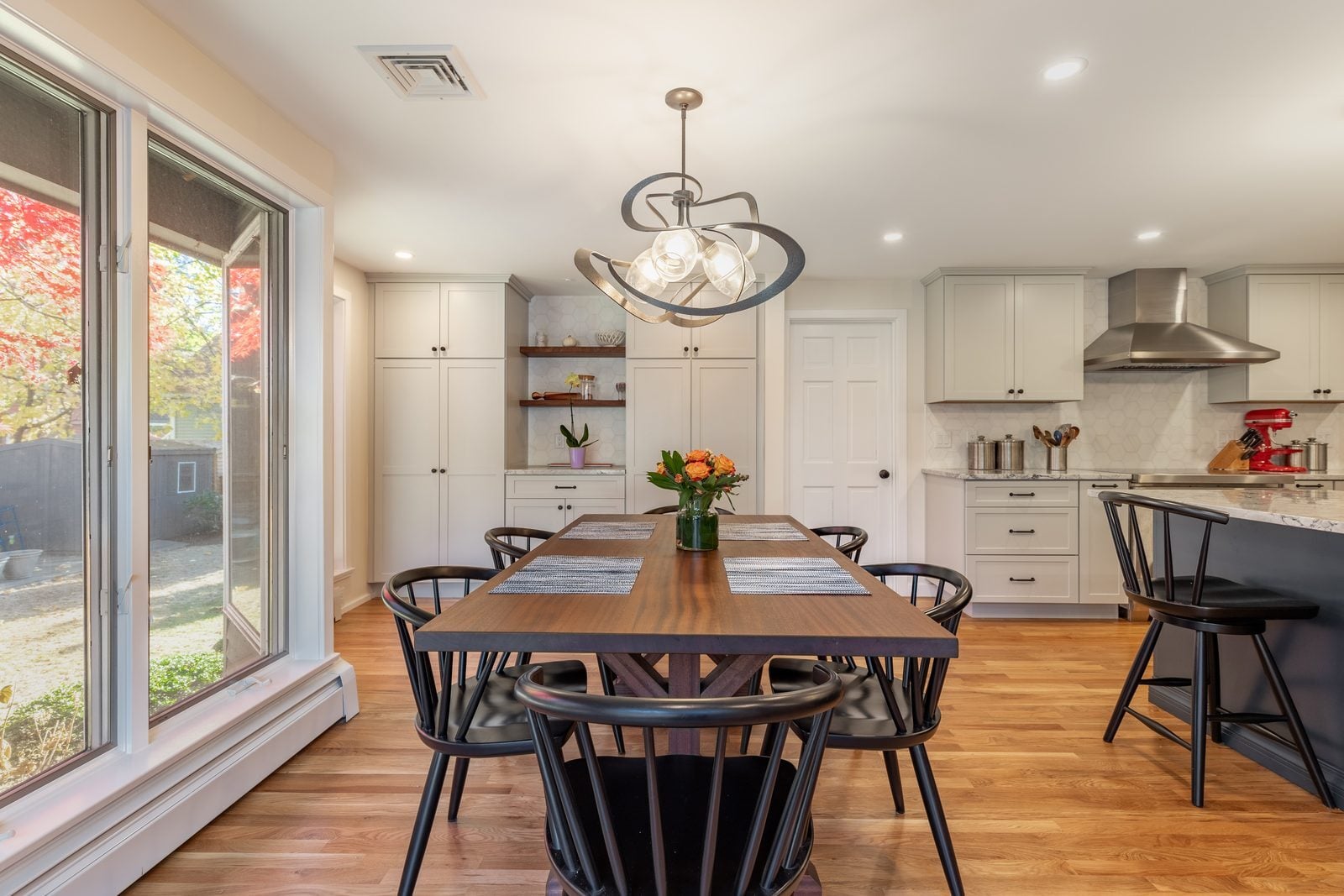 Dining table by tall windows with gray pantry cabinets, floating shelves and the range hood behind in Winchester, MA