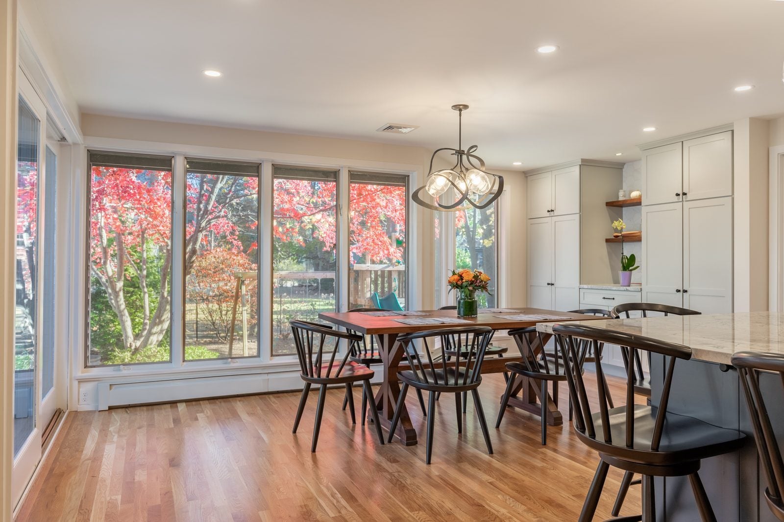 Dining area with a wall of windows facing fall trees, a trestle table and black spindle chairs next to the island in Winchester, MA