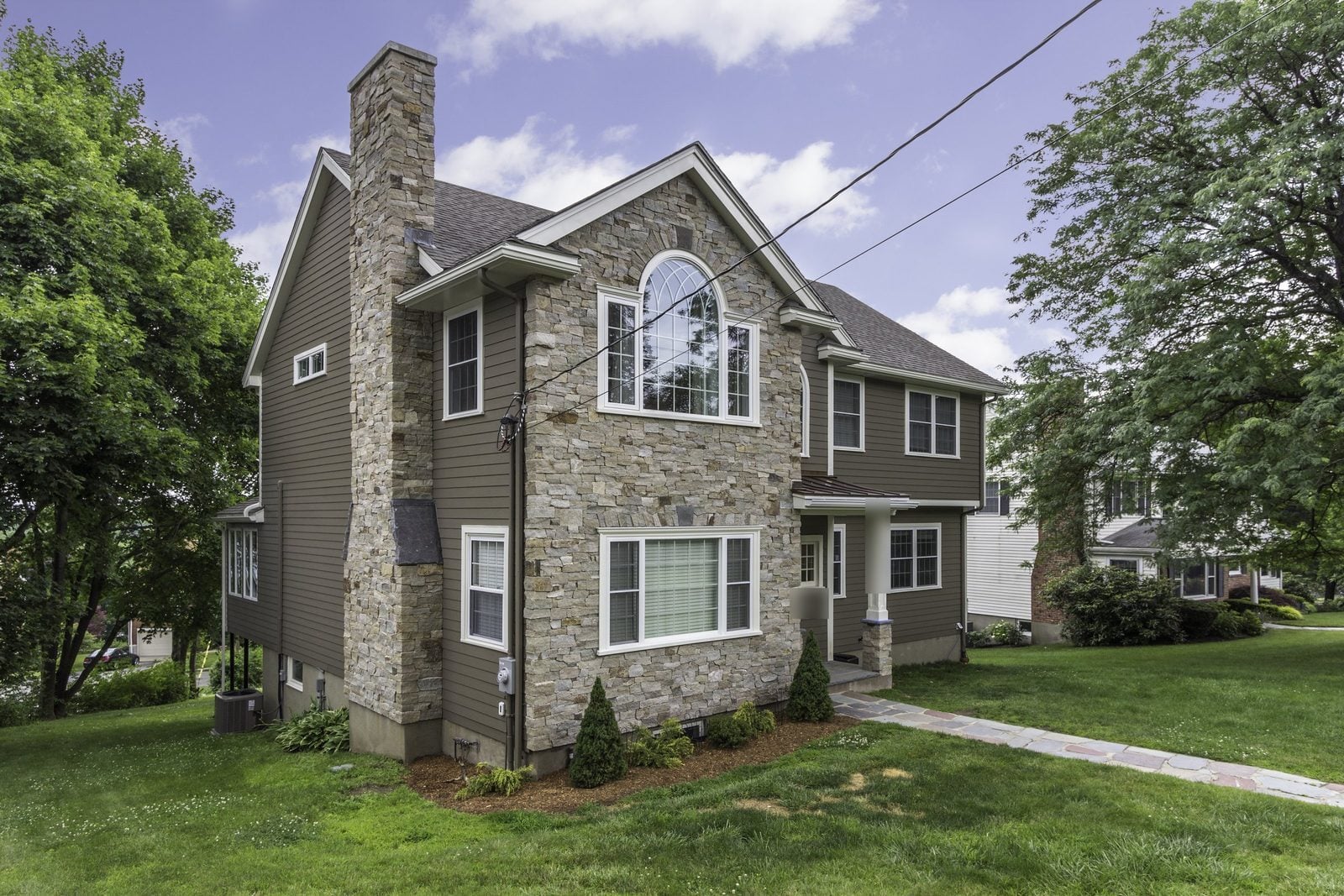 Corner view of a two-story home with stone chimney, stone-faced gable and brown siding in Winchester, MA